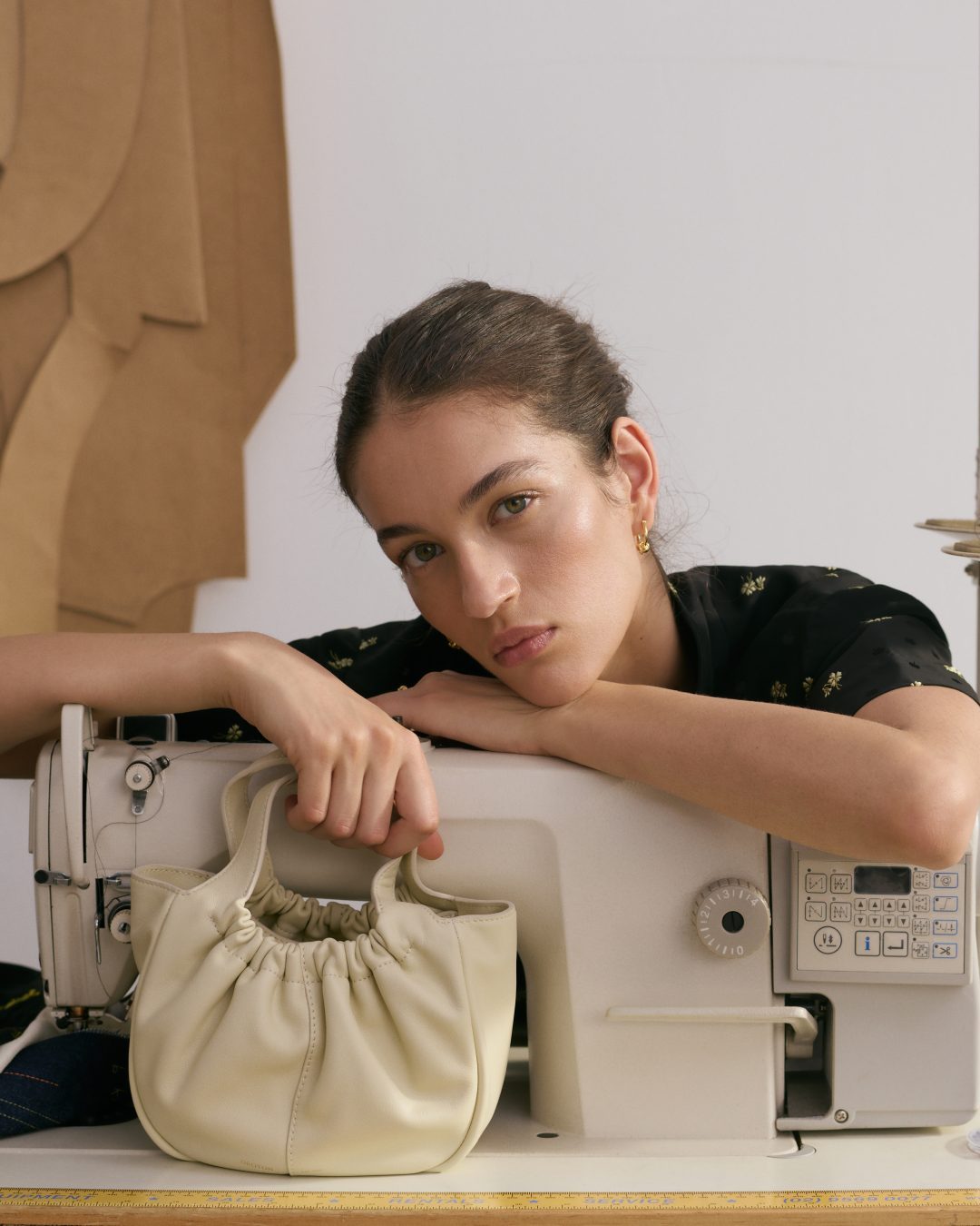 Woman rests on a sewing machine with a gathered-leather purse. The image showcases fashion, craftsmanship, and neutral tones.