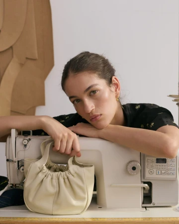 Woman rests on a sewing machine with a gathered-leather purse. The image showcases fashion, craftsmanship, and neutral tones.