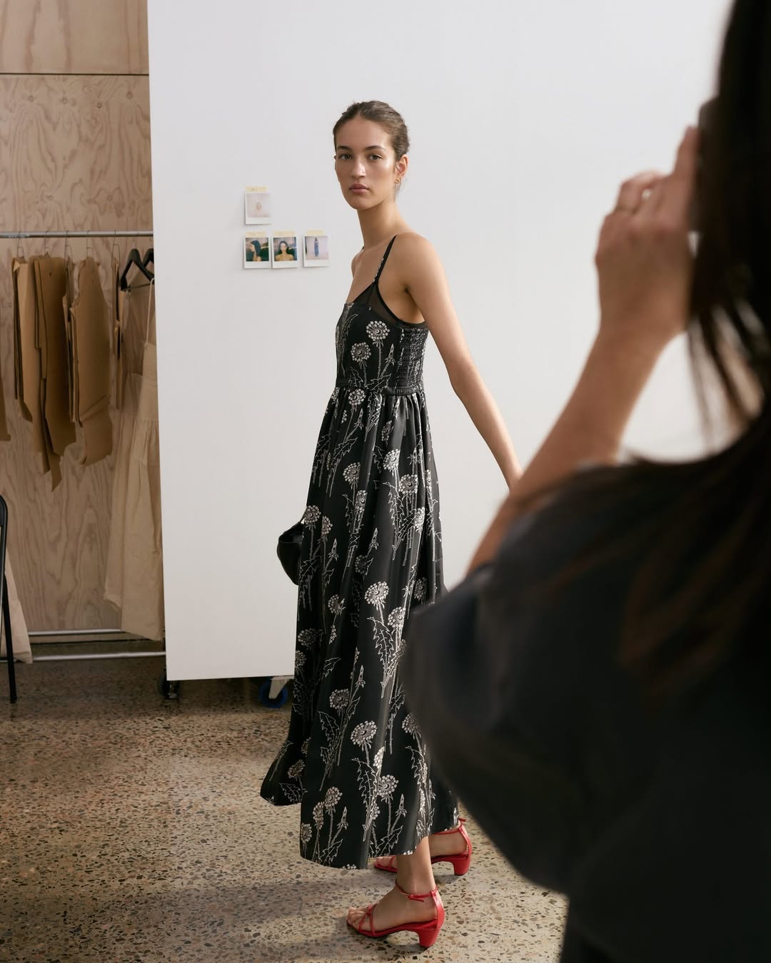Woman in black floral dress and red heels stands in a studio as a photographer takes her photo. Indoor fashion shot, medium framing.
