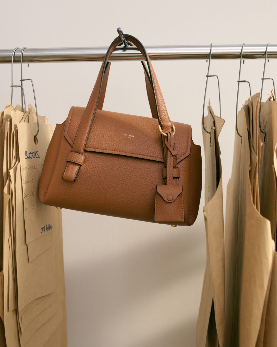 Tan leather handbag hanging on a metal garment rack alongside paper sewing patterns in a neutral, indoor setting.