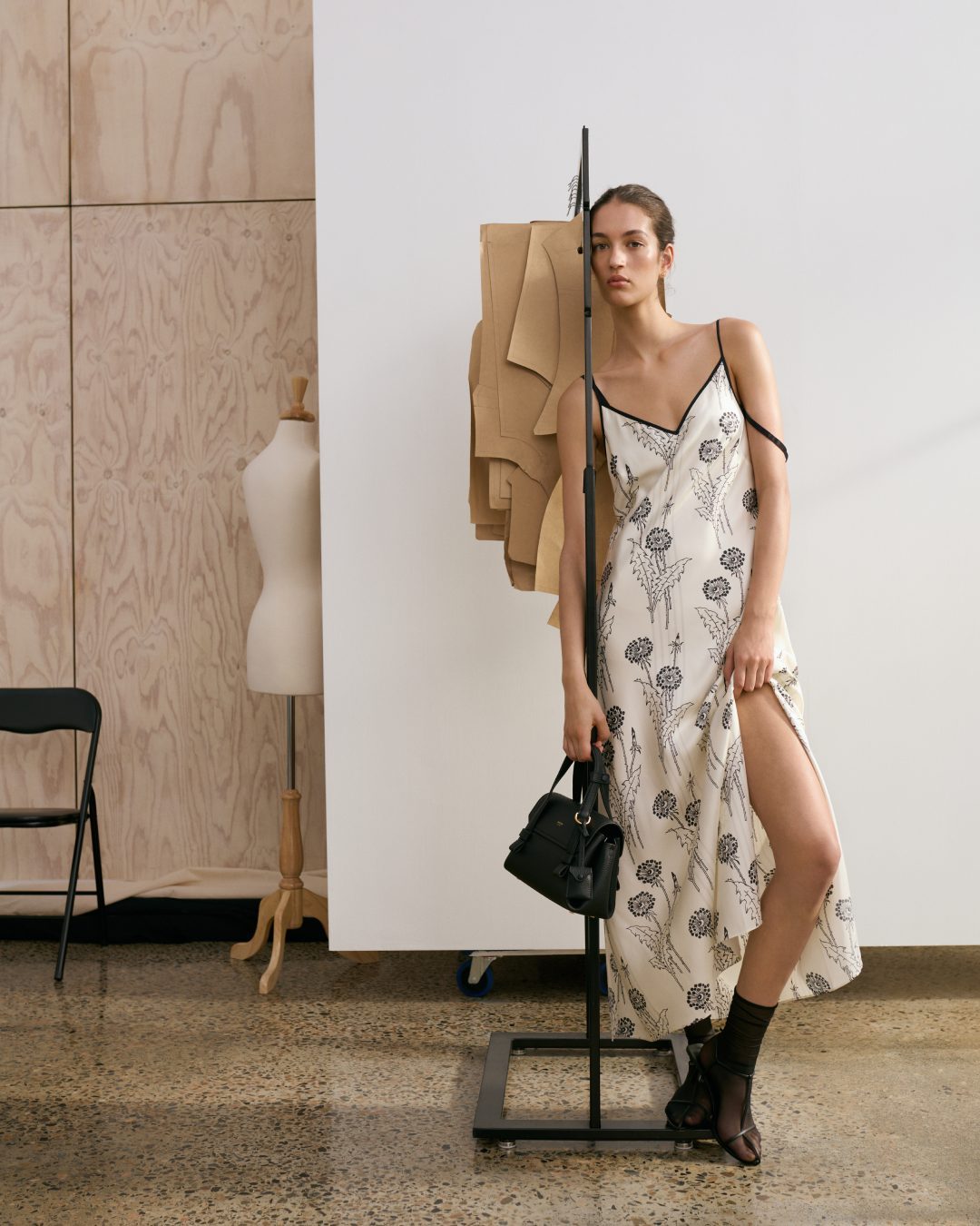 Woman poses indoors wearing a floral-print slip dress and sheer socks, holding a black bag. A dress form and sewing patterns are visible.