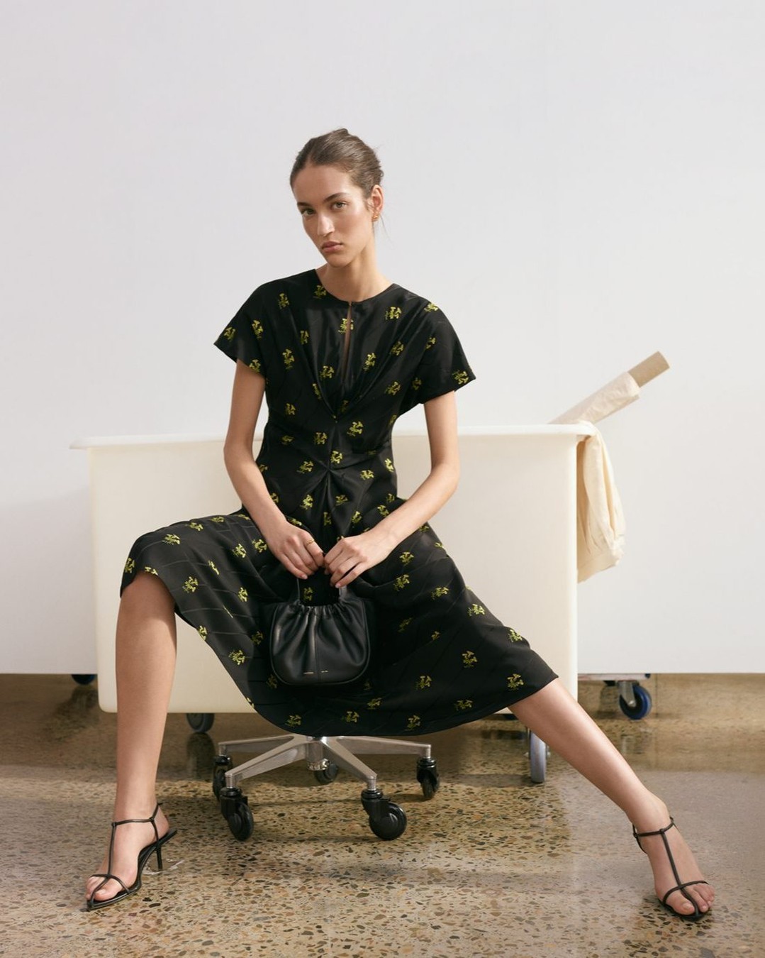 Elegant adult woman in a floral dress and heels sits on a rolling chair in front of a white desk holding a black purse in a studio.