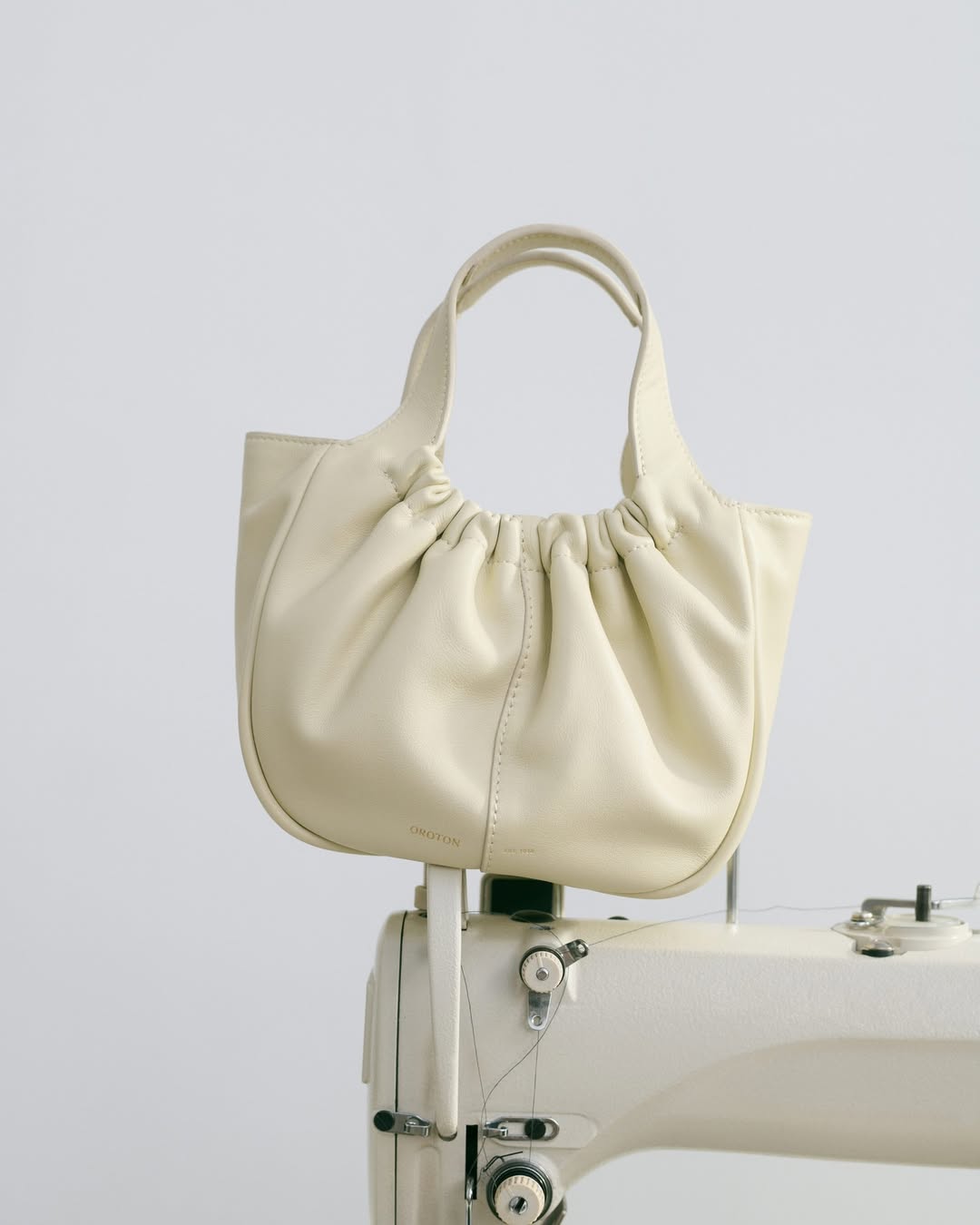 Cream-colored, gathered leather handbag with handles on a vintage white sewing machine against a neutral background.