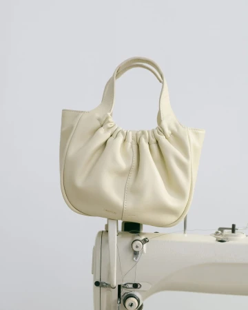 Cream-colored, gathered leather handbag with handles on a vintage white sewing machine against a neutral background.