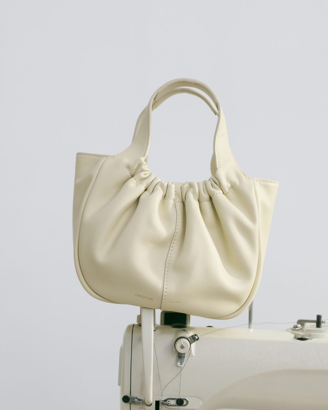Cream-colored gathered leather handbag with handles on a vintage sewing machine in a minimalist, neutral-toned product shot against white.