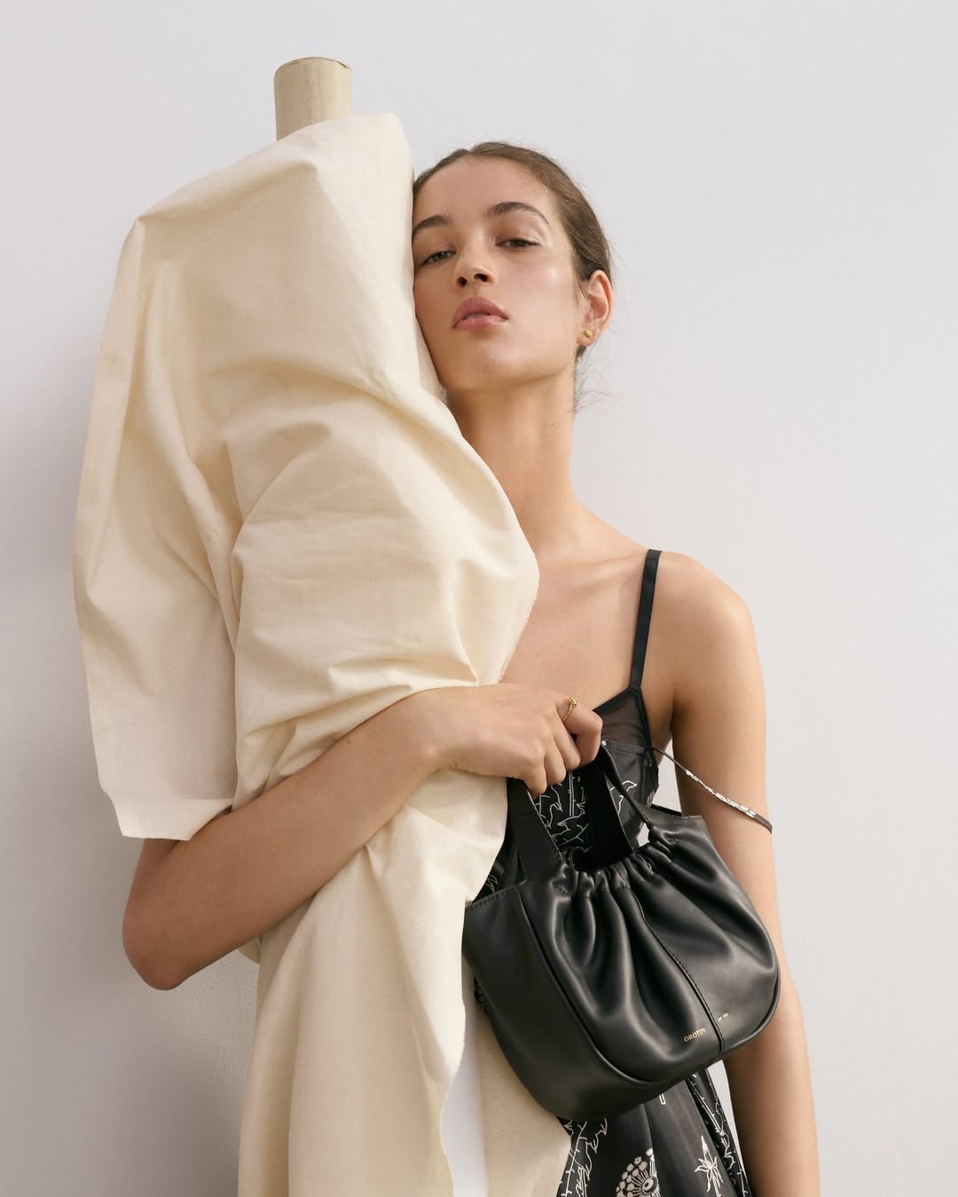 Adult woman poses in front of a mannequin with a black purse in a studio setting with diffused lighting for a fashion shoot.