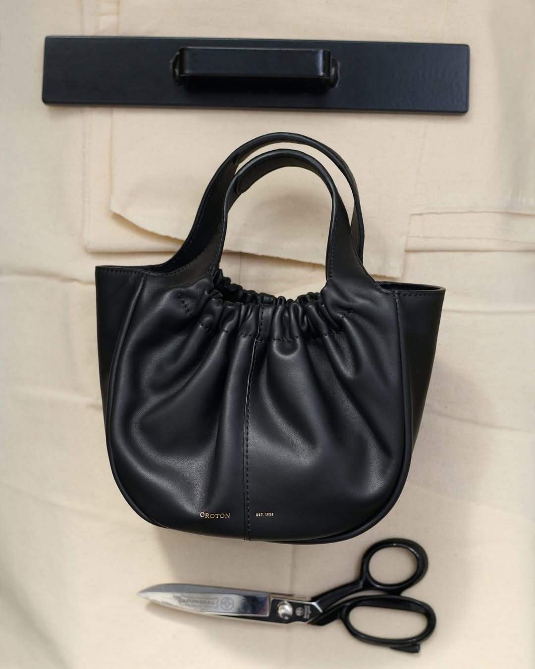Black leather ruched handbag with a metal bar handle, complemented by silver scissors and a neutral background.