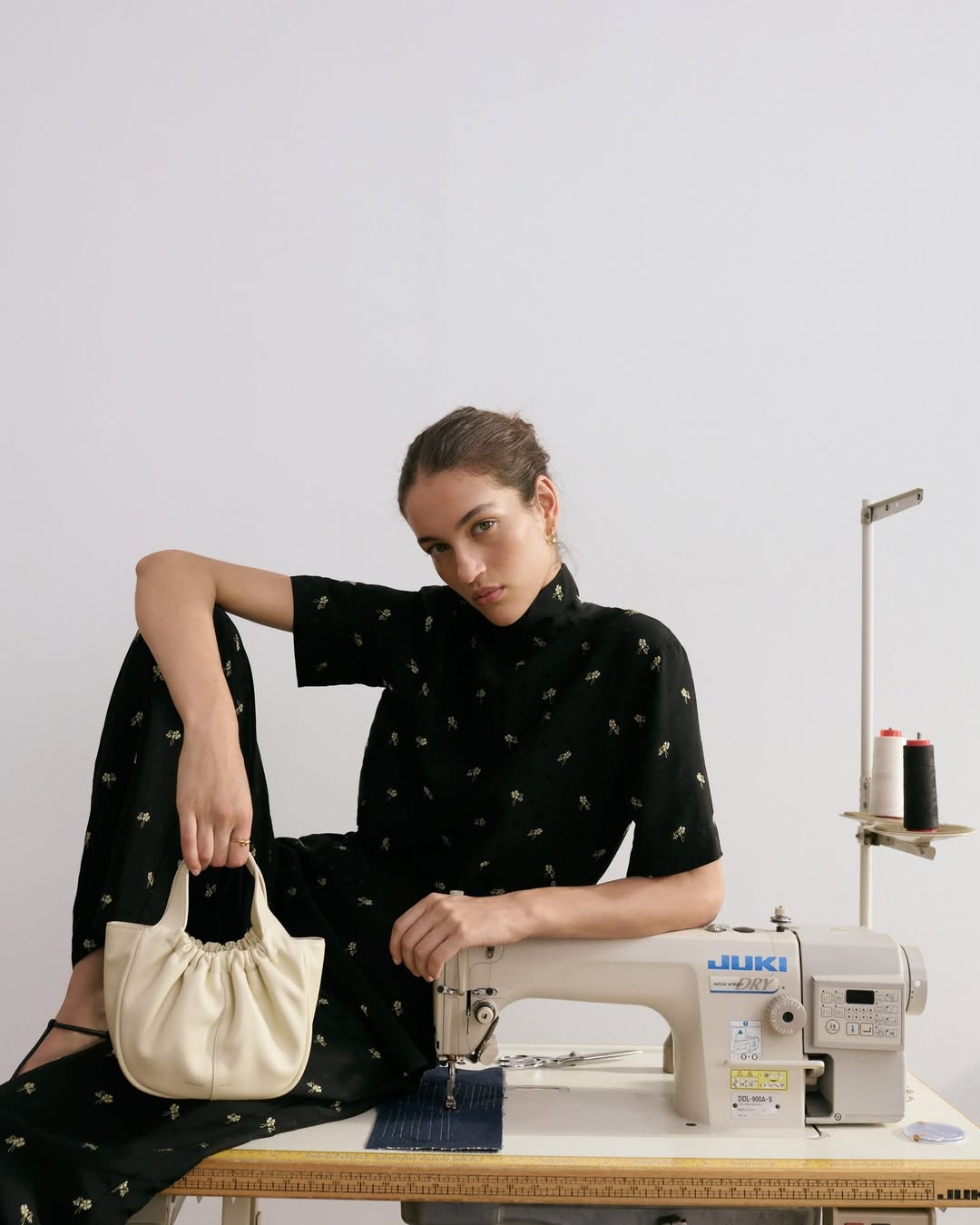 Fashion shot of a woman in a black dress holding a cream purse and posing next to a sewing machine on a white backdrop.