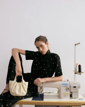 Fashion shot of a woman in a black dress holding a cream purse and posing next to a sewing machine on a white backdrop.