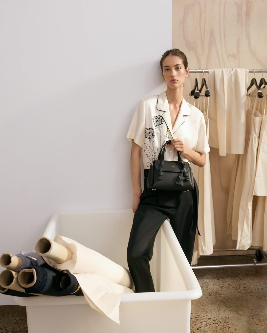 A woman poses with a black purse in a studio with fabric rolls and dresses, showcasing fashion and design elements in a clean setting.