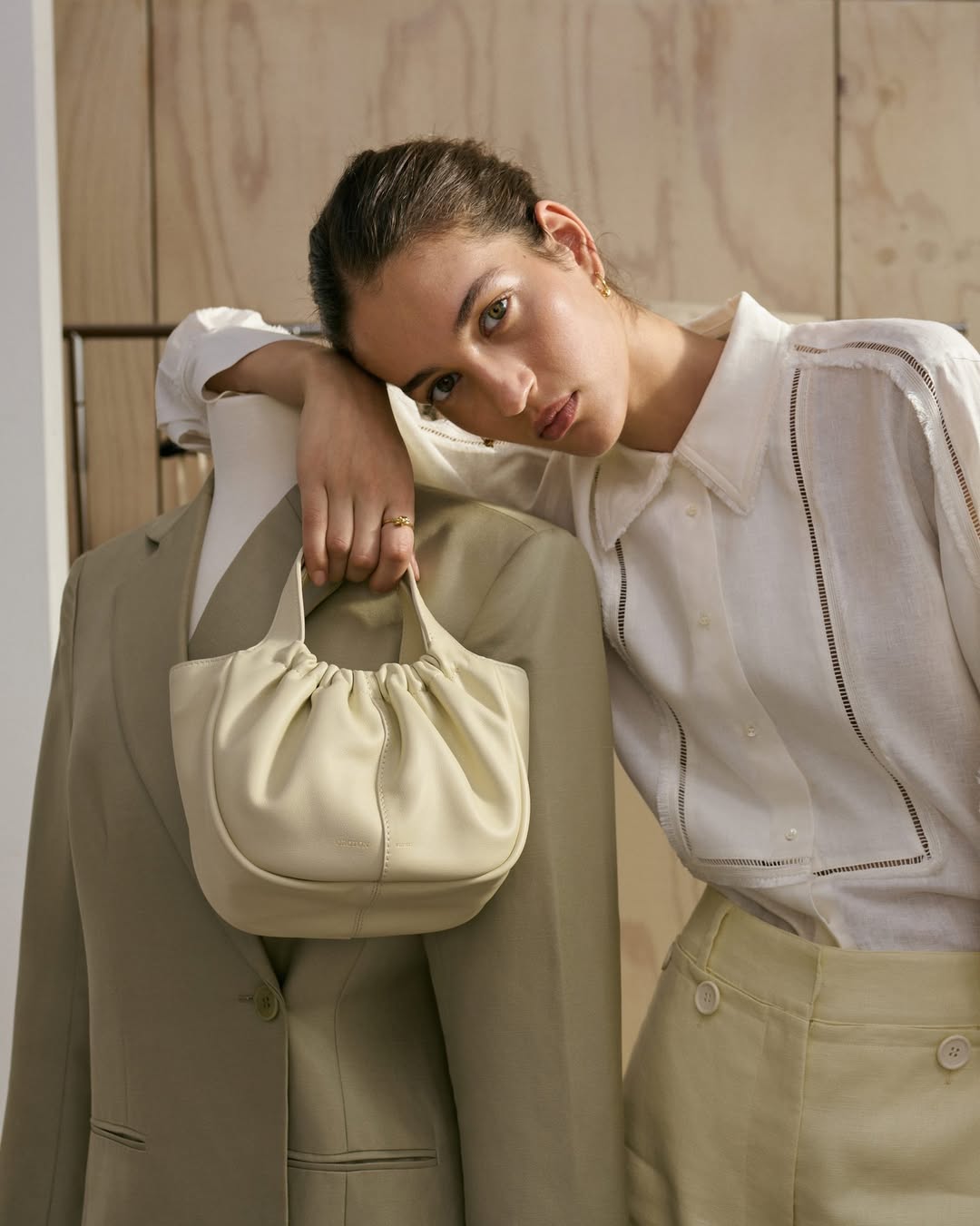 A woman leans against a mannequin wearing a blazer, holding a gathered leather purse against a wood paneled background in a fashion shot.