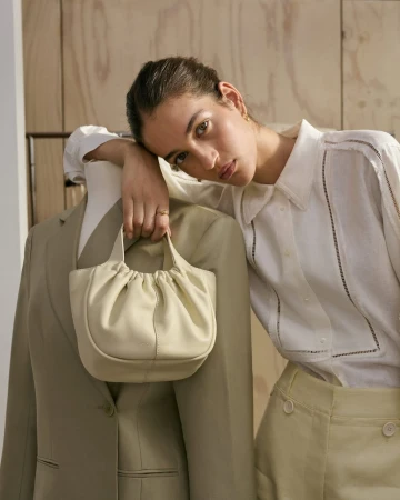 A woman leans against a mannequin wearing a blazer, holding a gathered leather purse against a wood paneled background in a fashion shot.