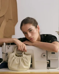 Model leaning on a sewing machine while holding a cream leather handbag