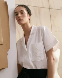 Model wearing a white short-sleeve shirt with lace pocket detail and black trousers against a neutral wall