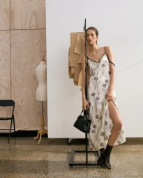 Model wearing a patterned slip dress holding a black handbag near a clothing rack in a studio setting