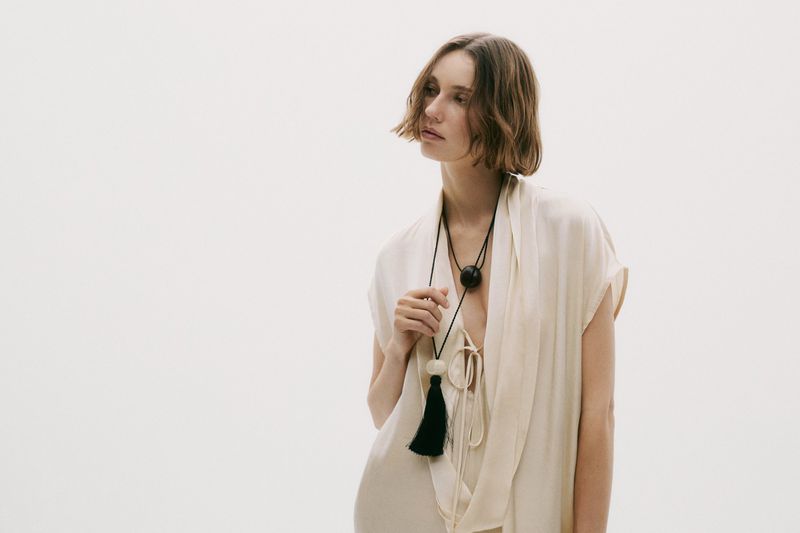 Woman in flowing white dress with a statement tassel necklace against a white backdrop in a fashion or beauty editorial style shot.