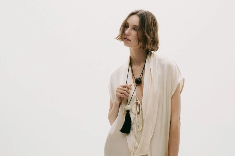 Woman in flowing white dress with a statement tassel necklace against a white backdrop in a fashion or beauty editorial style shot.