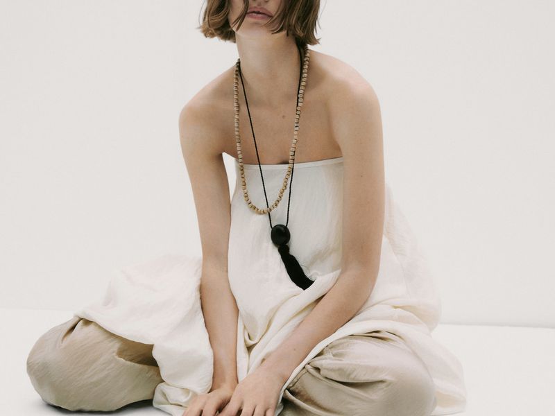 A woman wearing a long beaded necklace sits with her hands clasped in a neutral cream-colored, strapless, loose fitting dress against white.