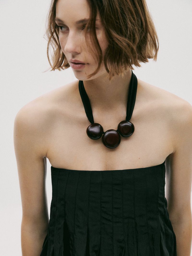 Young woman with short hair wears a strapless black dress and a chunky wooden necklace in a minimalist studio setting.