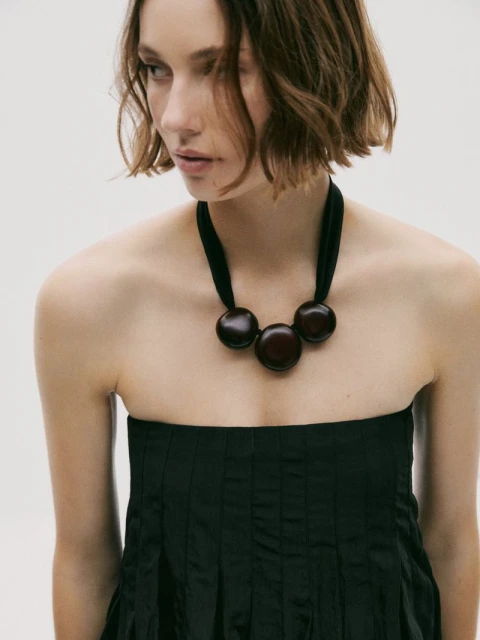 Young woman with short hair wears a strapless black dress and a chunky wooden necklace in a minimalist studio setting.