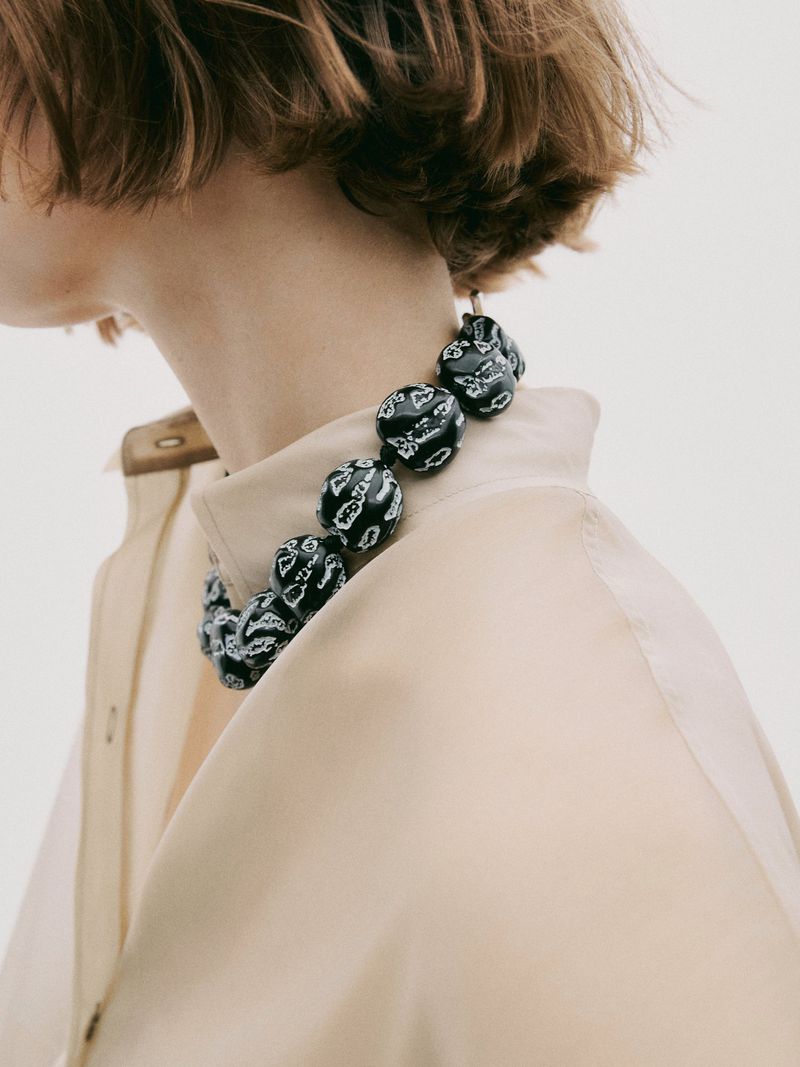 Adult woman wearing a black beaded necklace and beige collared jacket, photographed in a brightly lit photo studio.