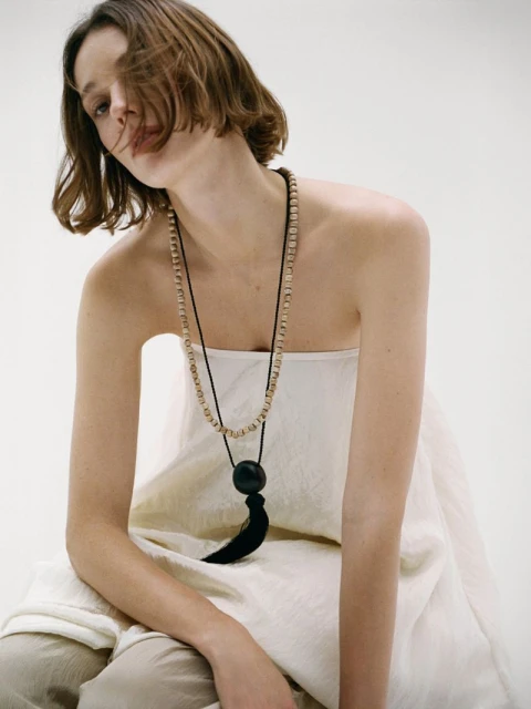 Adult female model poses in a studio wearing a long beaded necklace with a large black tassel. Soft neutral lighting. Fashion shot.
