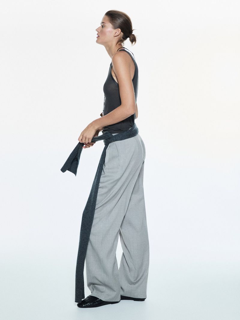 A woman poses in a gray tank top and pinstripe pants, cinched with a long gray scarf, in a studio setting against a white backdrop.