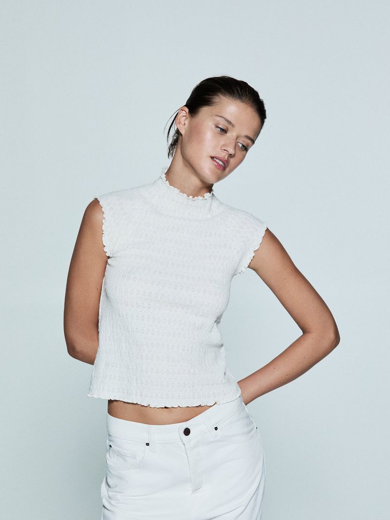 A young adult woman poses in a studio wearing a white textured short sleeve top and white jeans, hands on hips, against a neutral backdrop.