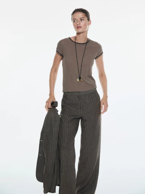 Fashion shot of a slender adult woman in pinstripe pants and a brown t-shirt holding a jacket, posing in front of a white background.