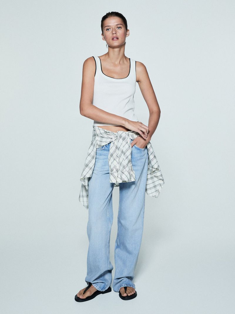 Woman in a white tank top and light wash jeans in a studio. Flannel shirt tied around her waist. Fashion lifestyle photo.