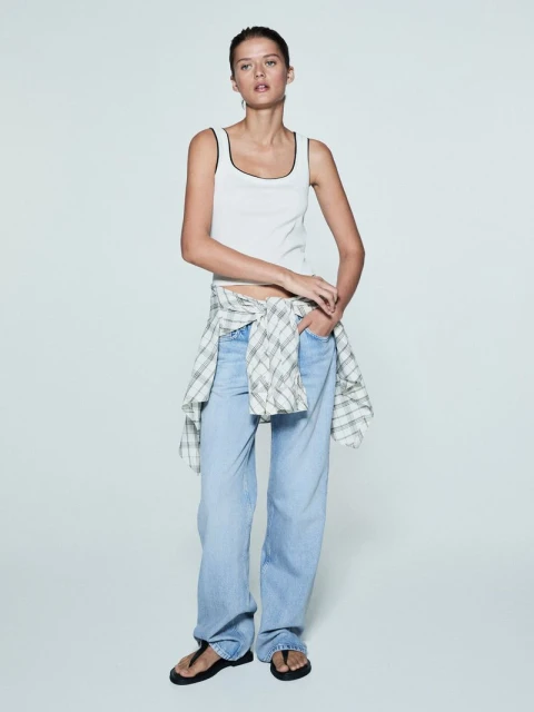 Woman in a white tank top and light wash jeans in a studio. Flannel shirt tied around her waist. Fashion lifestyle photo.
