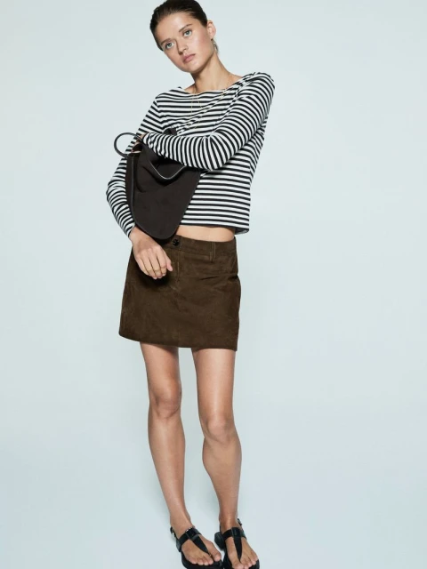 Fashionable woman in a striped top and brown skirt poses in a studio, showcasing a casual yet chic style with a modern aesthetic.