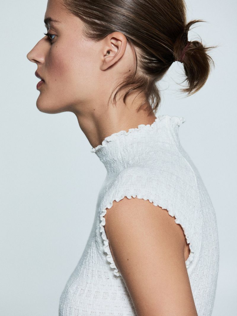 Profile portrait of a woman with dark hair pulled back, wearing a textured white top with a ruffled neckline against a white background.