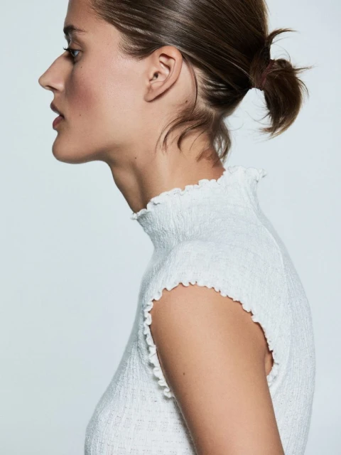 Profile portrait of a woman with dark hair pulled back, wearing a textured white top with a ruffled neckline against a white background.
