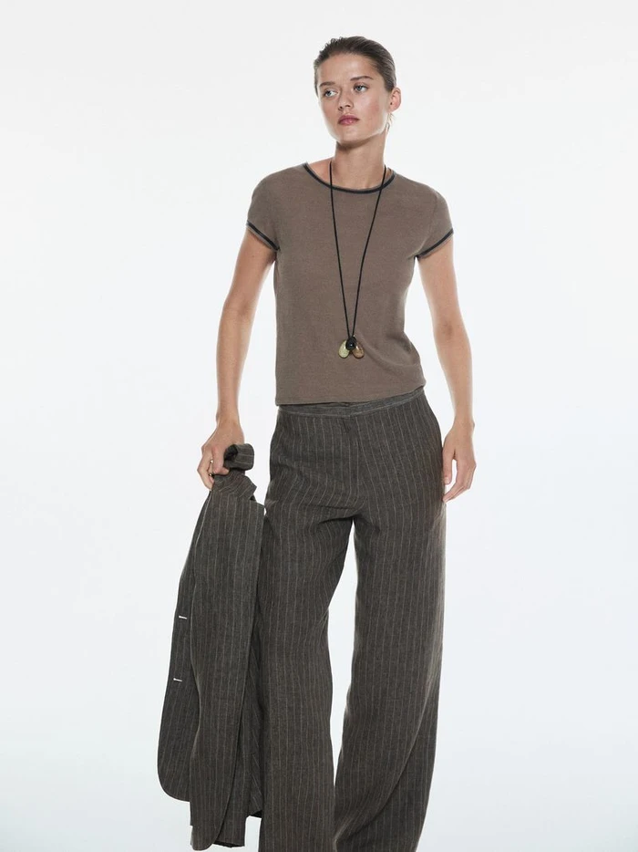 Model wearing a brown t-shirt and striped trousers against a plain white background holding a dark jacket