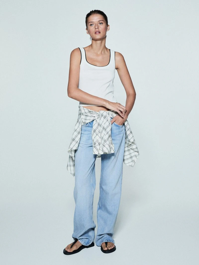 Model wearing a white tank top, light blue jeans, and a plaid shirt tied around her waist against a solid grey background