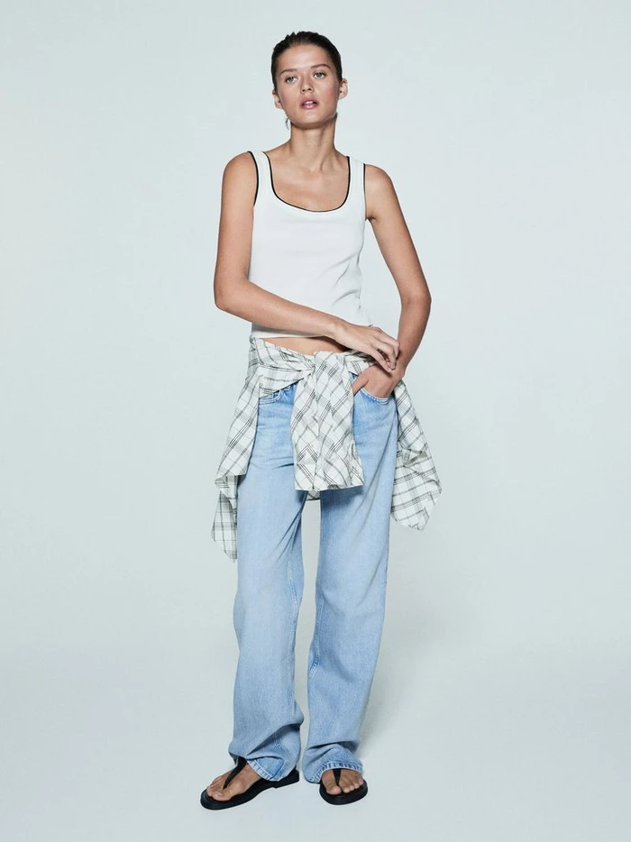 Model wearing a white tank top, light blue jeans, and a plaid shirt tied around her waist against a solid grey background