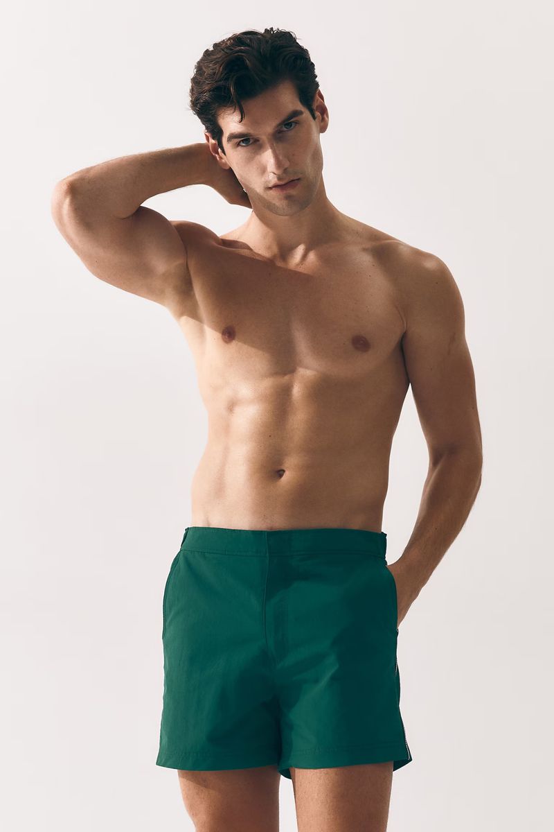 Shirtless man in green shorts poses in a studio with hand behind head. Head and shoulders shot, lit from above with soft focus.