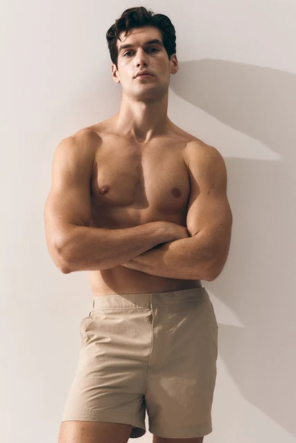 Athletic man in khaki shorts posing indoors with arms crossed in front of a cream-colored wall, lit with soft, natural-looking light.