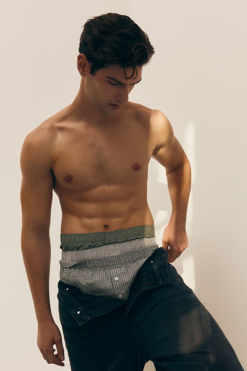 Young adult male in underwear and jeans standing in a studio with soft lighting, looking down, showcasing menswear fashion.