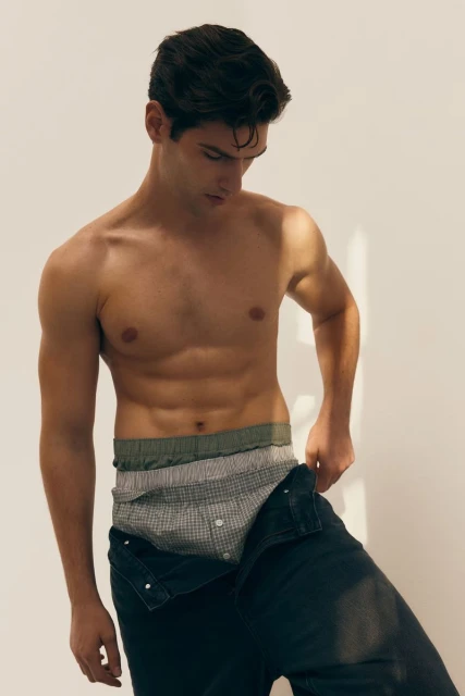 Young adult male in underwear and jeans standing in a studio with soft lighting, looking down, showcasing menswear fashion.