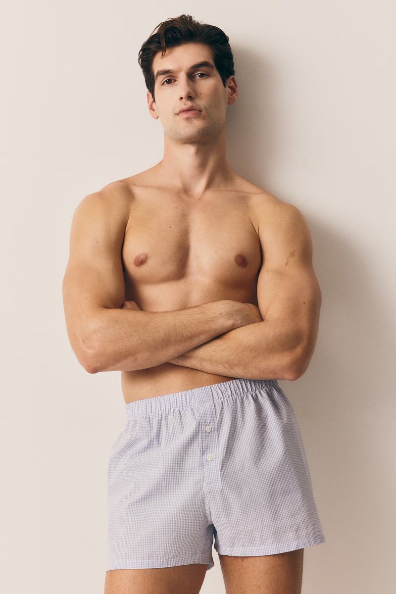 Athletic man in boxer shorts poses against a neutral wall. Upper body is bare with arms crossed, displaying casual comfort in indoor setting.