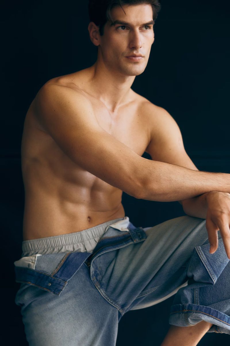 Adult male model poses shirtless in unbuttoned denim pants against a dark backdrop in a fashion forward portrait in a studio setting.