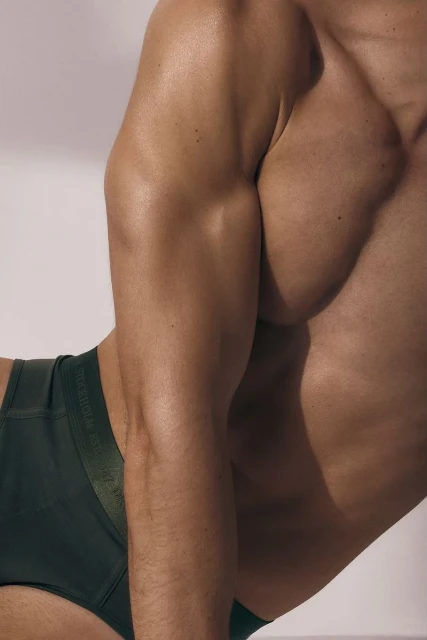 Close-up shot of a tanned adult male torso and arm against a white background, wearing dark green underwear with a logo. 