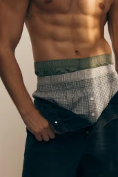 Close-up portrait of a male model wearing layered underwear and unbuttoned trousers against a clean background.