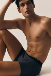 Shirtless male model wearing dark boxers seated against a clean white studio background for an H&M campaign.