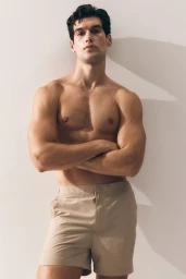 Shirtless male model with arms crossed wearing beige shorts against a white studio background.