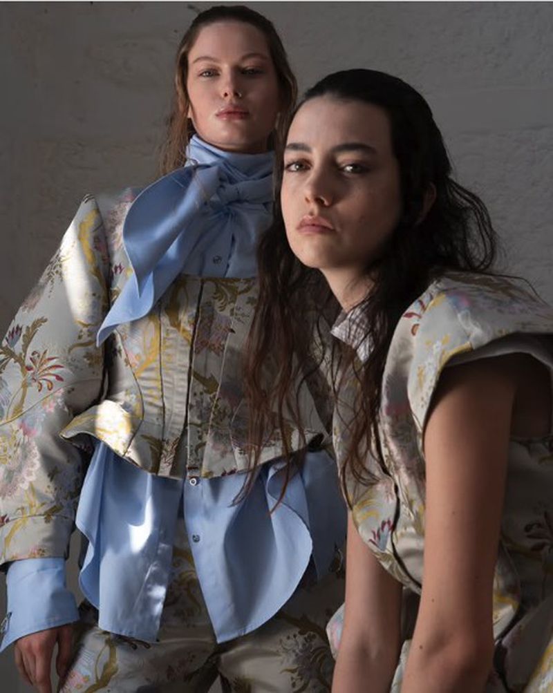 Two women model floral jackets and shirts in a studio setting with natural lighting, showcasing unique fashion apparel.