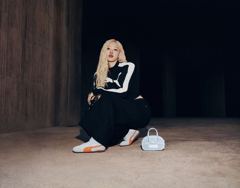 A blonde woman poses indoors wearing a Puma sweatshirt, black pants, and Puma sneakers with a Puma purse on the floor. 