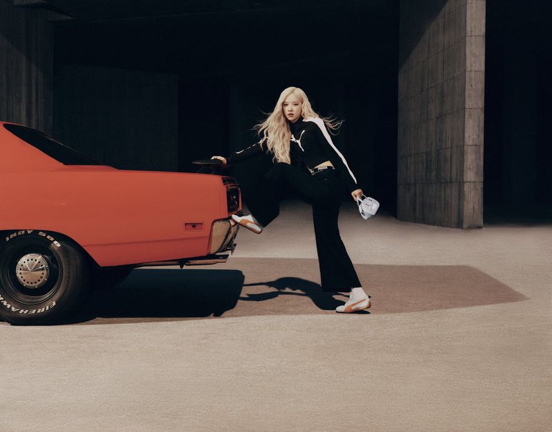 Stylish woman with long blonde hair poses with a vintage orange muscle car against a concrete wall in a fashion editorial photoshoot.